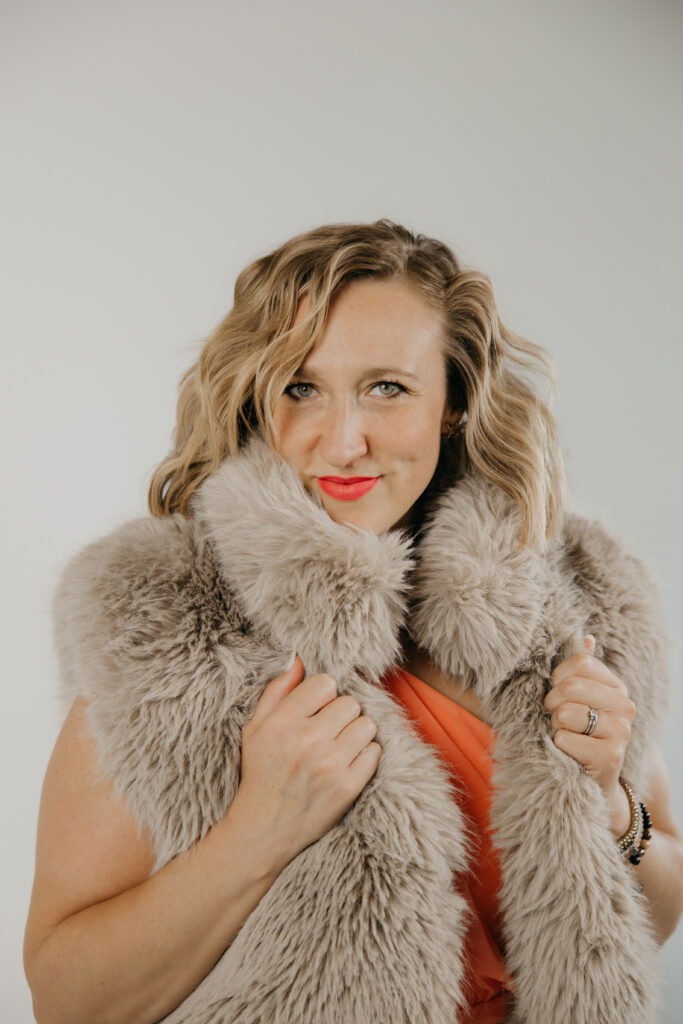 Close-up portrait of Dr. Mel Krug in a fur stole over an orange dress, red lipstick, grey backdrop