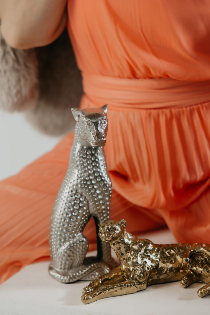 Cropped shot of an orange dress and hand resting near gold and silver leopard statues, brand photography styling detail