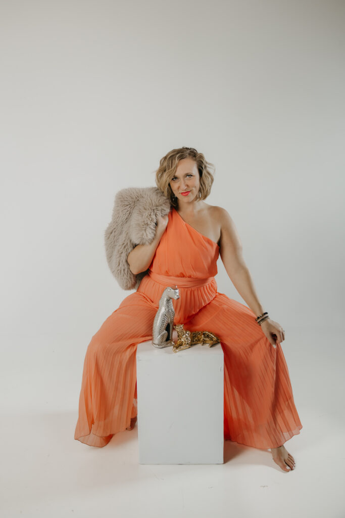 Dr. Mel Krug seated on a white pedestal with a fur stole draped over one shoulder, orange dress and leopard statues