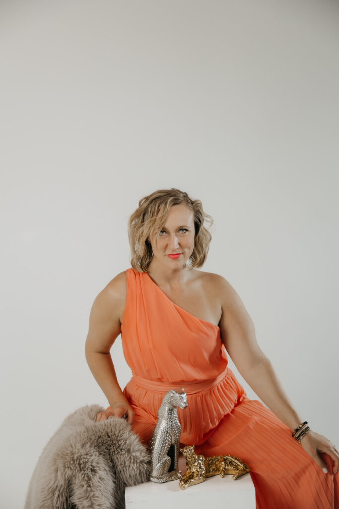 Direct-gaze portrait of Dr. Mel Krug seated on a white pedestal in a fur stole and orange dress