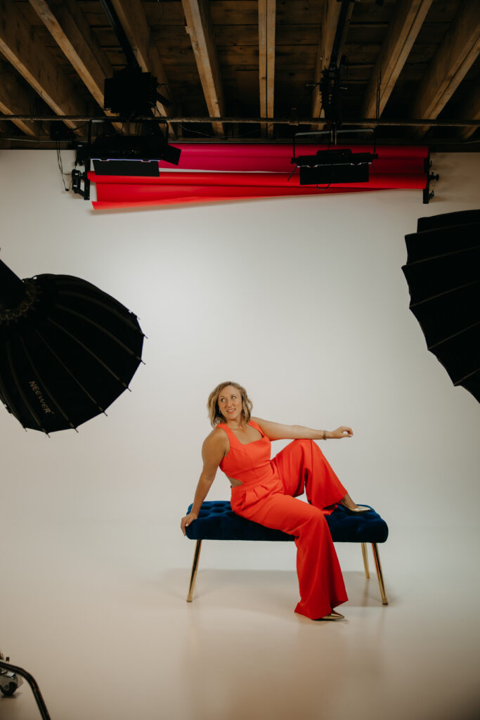 Wide shot of a woman seated on a blue velvet bench with studio softbox lighting visible