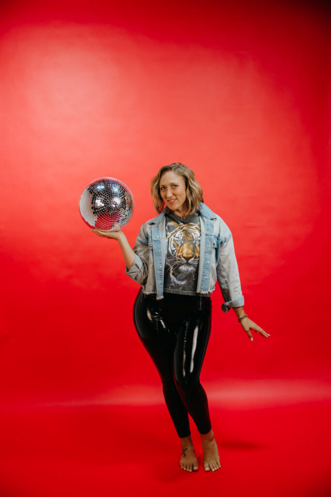 Woman standing and holding a single disco ball, playful pose against a red backdrop