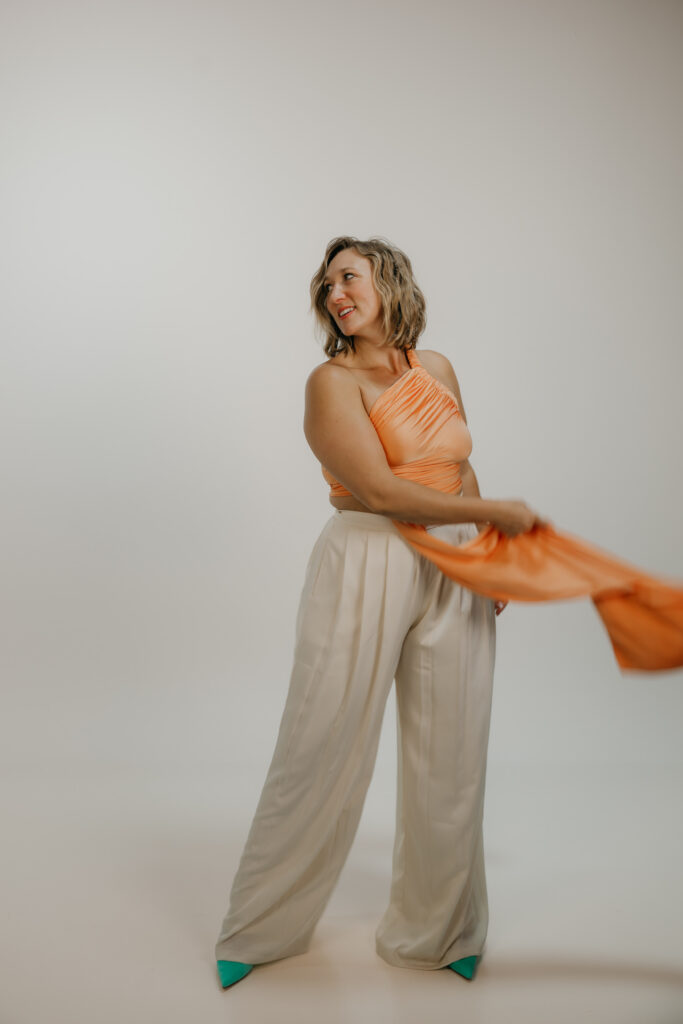 Woman pulling orange fabric to the side in a playful movement, white trousers