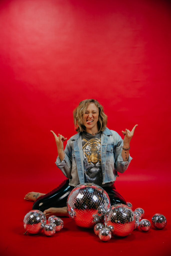Woman seated cross-legged making rock hand gestures with both hands, disco balls scattered around her on a red backdrop