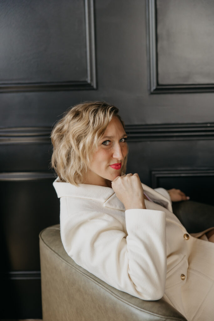 Transformational coach reclined in a dark leather chair against a black paneled wall, cream blazer brand portrait