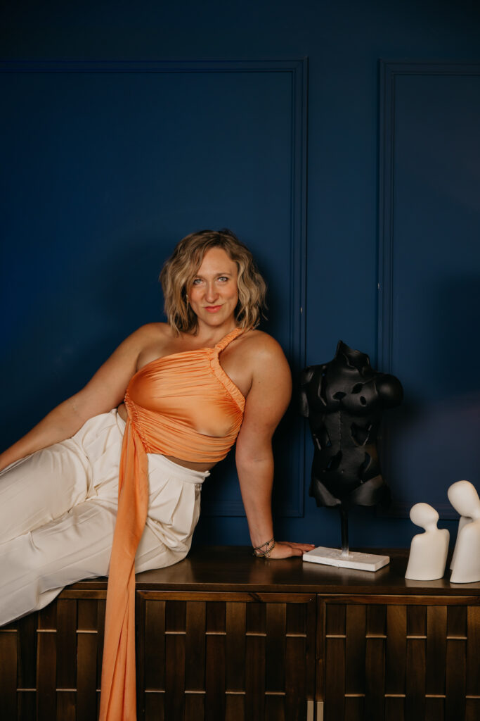 Woman reclined on a wood credenza in an orange wrap top beside ceramic decor pieces