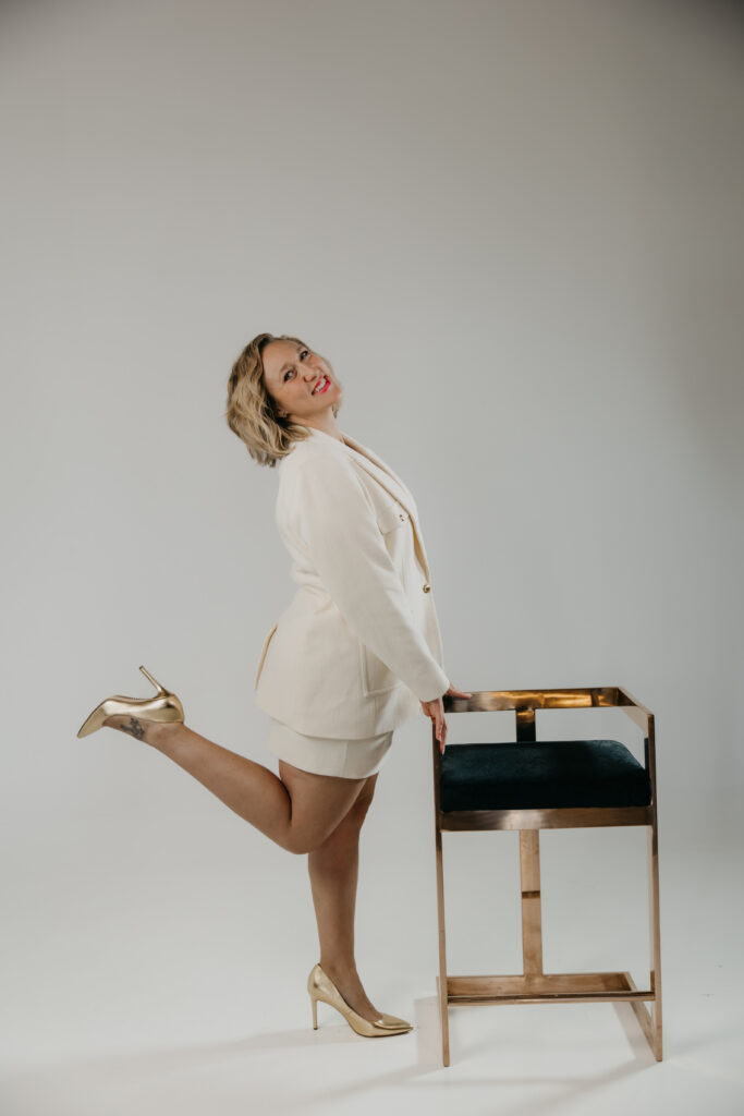 Playful brand photography pose, one leg raised on a gold stool, cream blazer suit