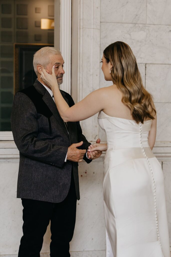 Bride's father touches her face during their first look on a marble staircase