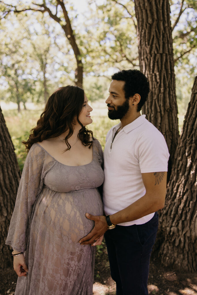 Emily and Calvin standing close together, hands on her belly, framed by a large oak trunk