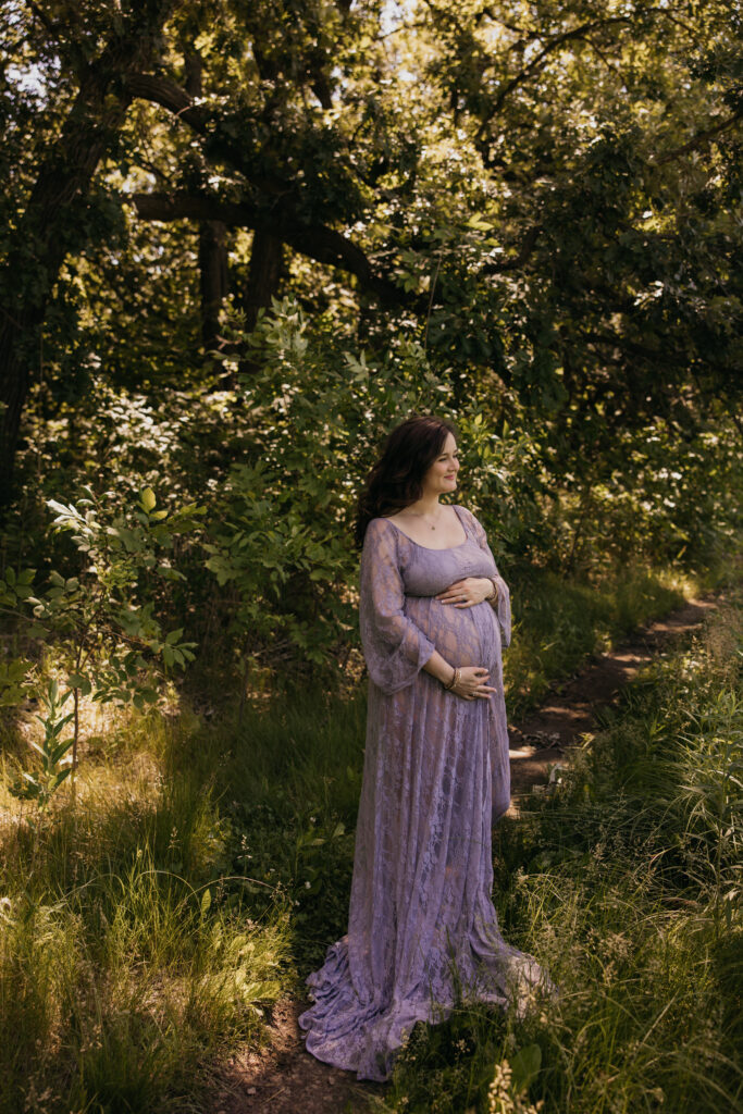 Emily looking down thoughtfully at her belly, standing on a sunlit forest path
