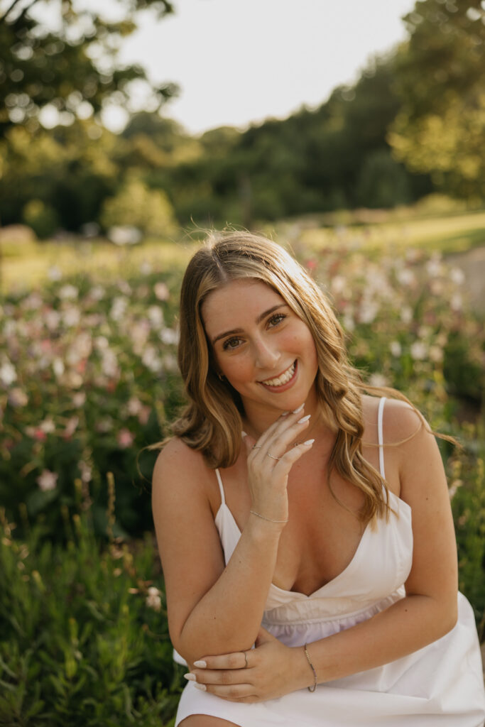 senior girl in white dress sitting close-up, hand near face, pink flowers behind her