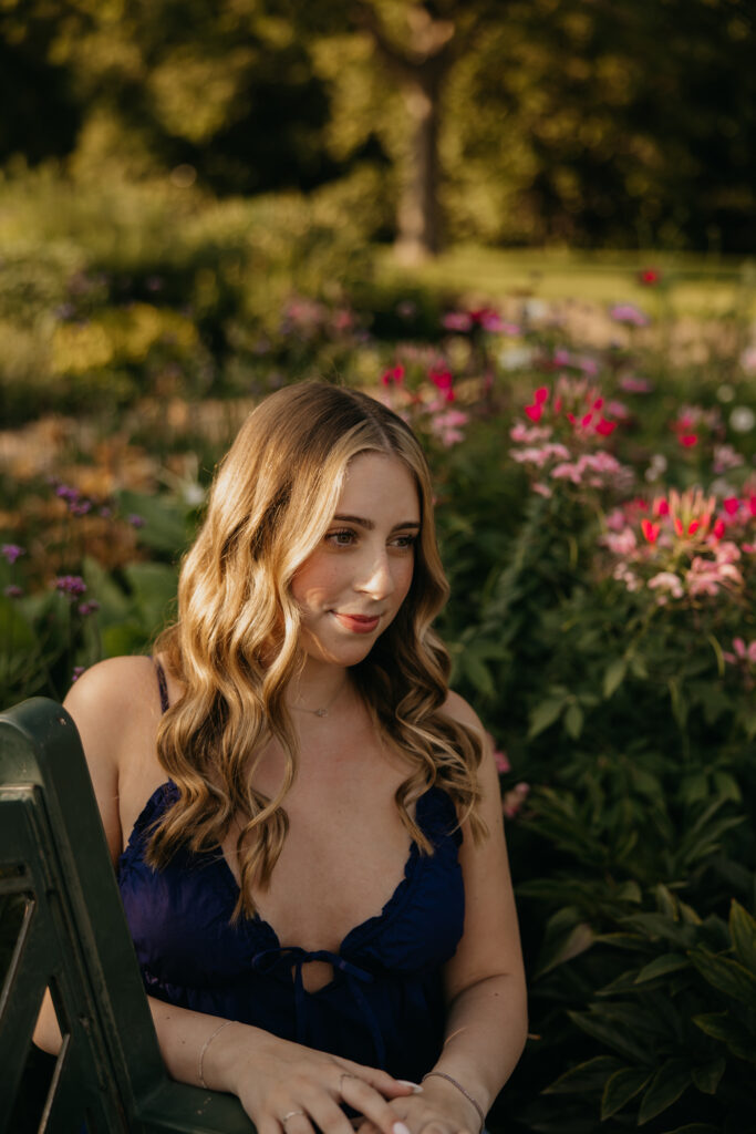 senior girl in navy top sitting on garden bench, looking down among pink flowers