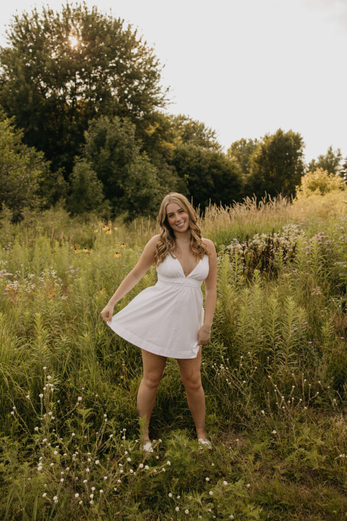 senior girl in white dress standing in wildflower field, holding dress out at sides