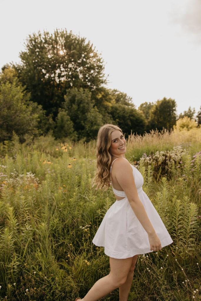 senior girl in white dress walking through tall grass, dress caught mid-motion
