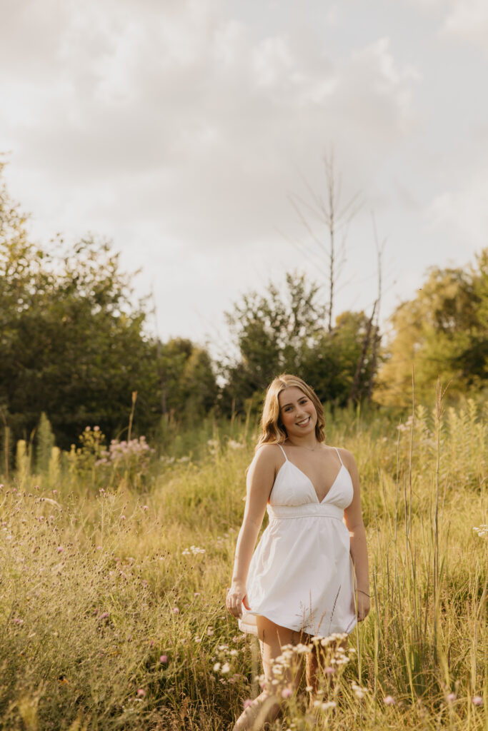 senior girl in white dress standing in field, looking down as dress sways in the breeze