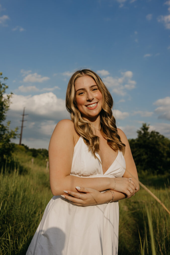 senior girl in white dress standing with arms crossed, blue sky background