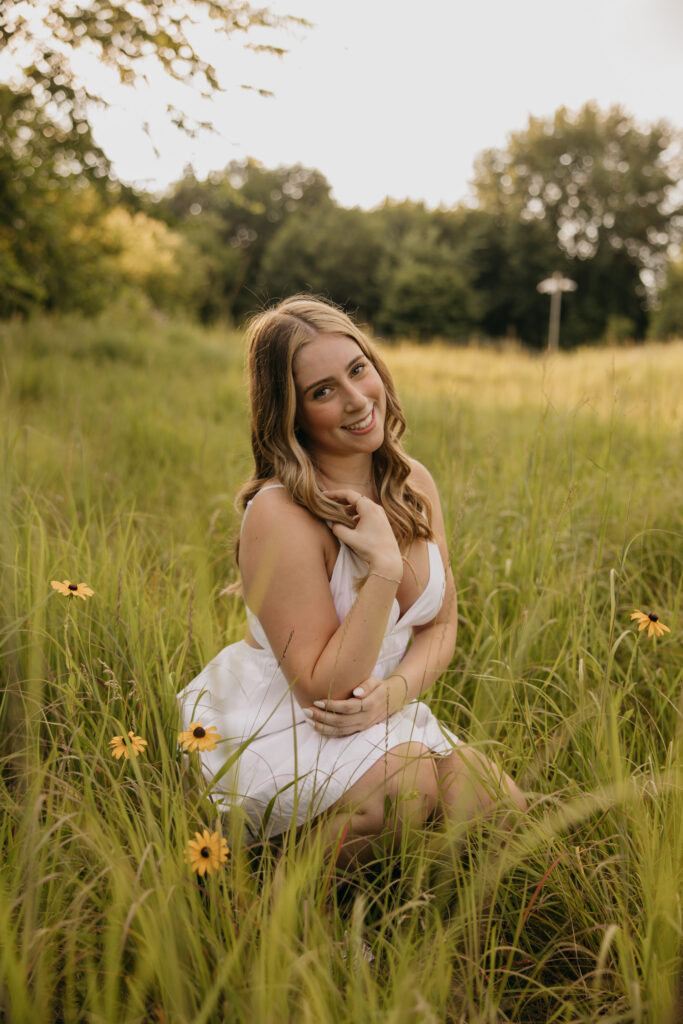 senior girl in white dress crouching in wildflower field, smiling with hand near chin