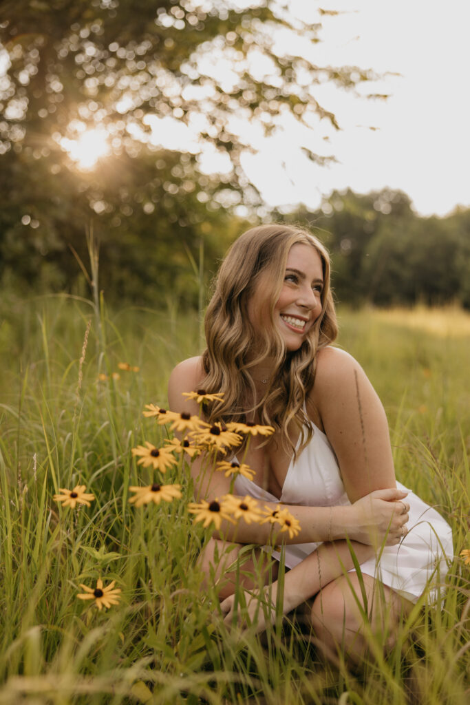 senior girl in white dress crouching in tall grass, laughing softly