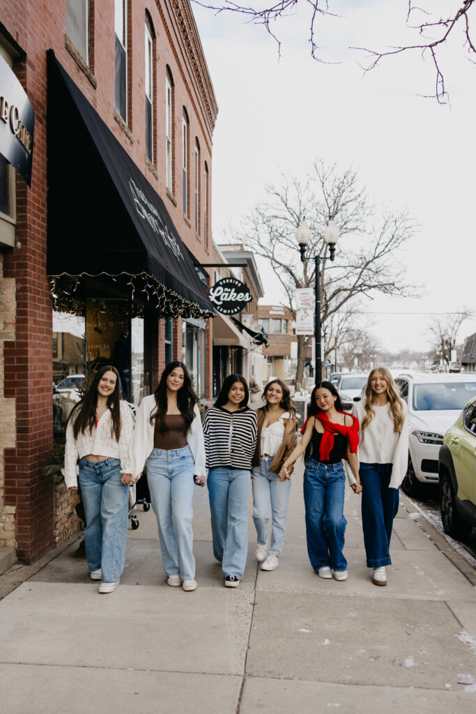 Six senior friends in cream, brown, and red walking together past a brick building with a black awning