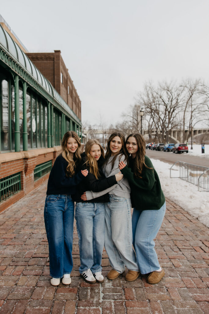 Four senior friends in coordinating sweaters standing arm in arm on a brick sidewalk beside a glass storefront