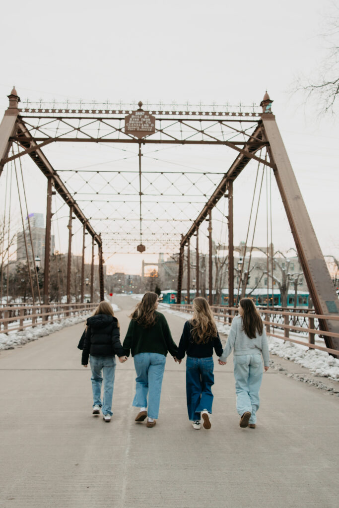 Four senior friends walking hand in hand away from the camera across a truss bridge at sunset