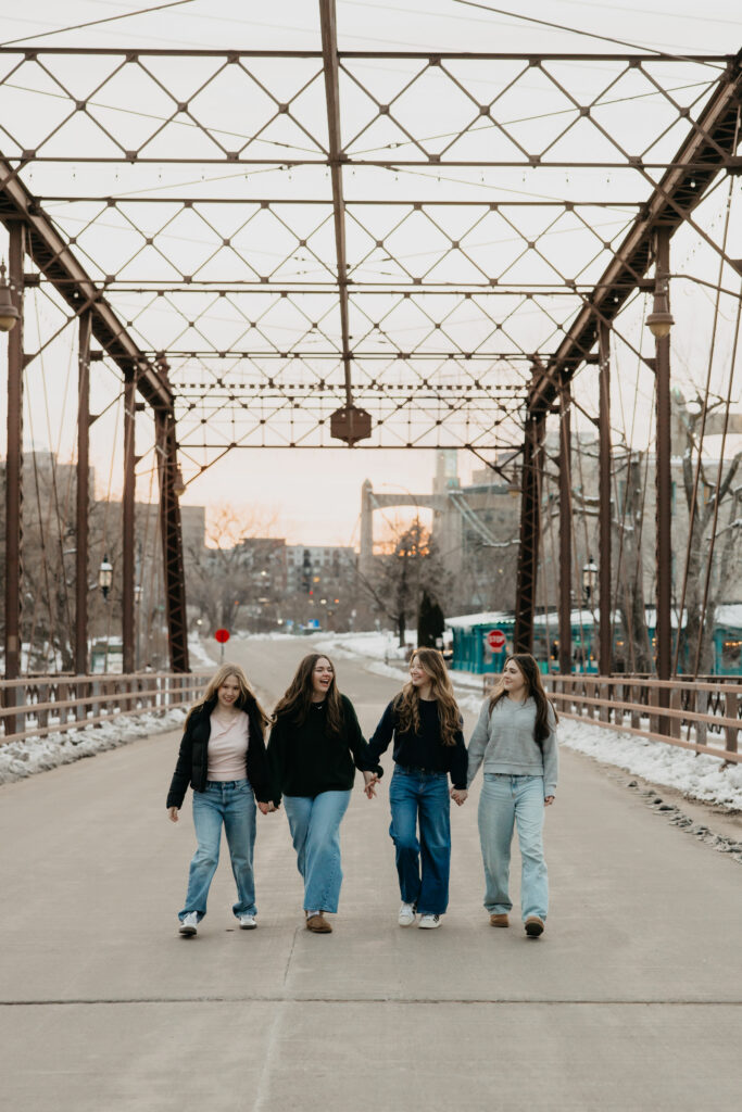 Four senior friends walking hand in hand toward the camera across a Minneapolis pedestrian bridge