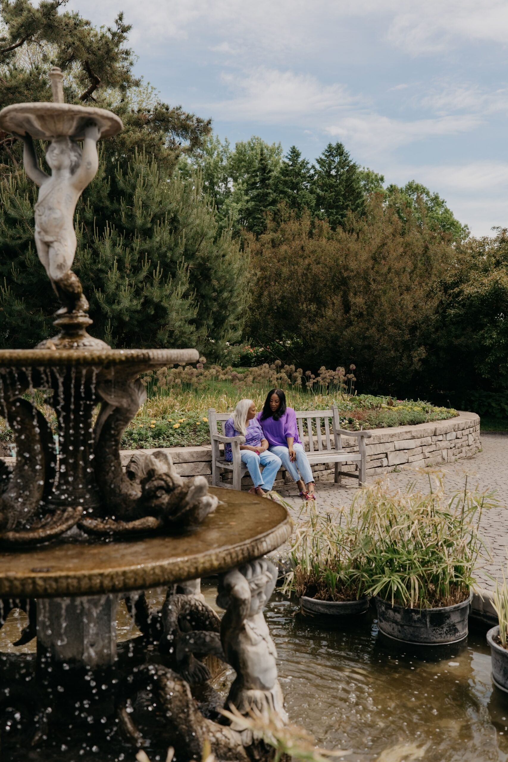 Mother and daughter in mother daughter matching outfits sitting on a garden bench beside a stone fountain