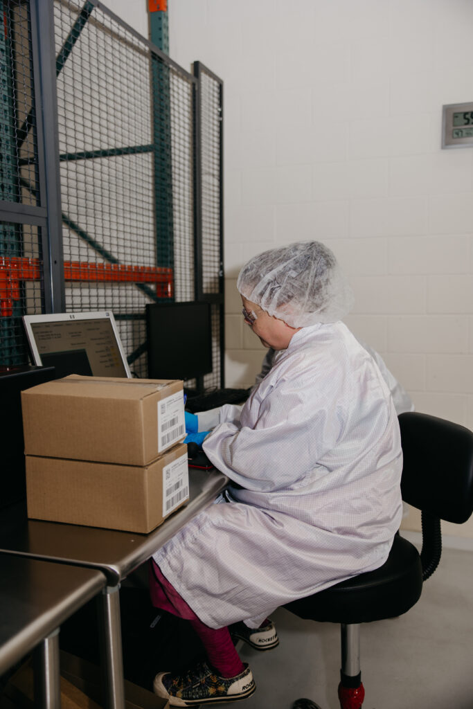 Technician in white gown processing packaged product boxes at a warehouse workstation
