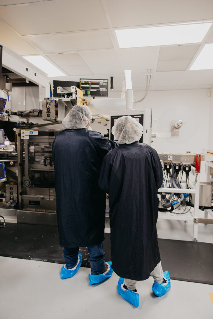 Two technicians in dark gowns and blue shoe covers reviewing manufacturing equipment monitors together