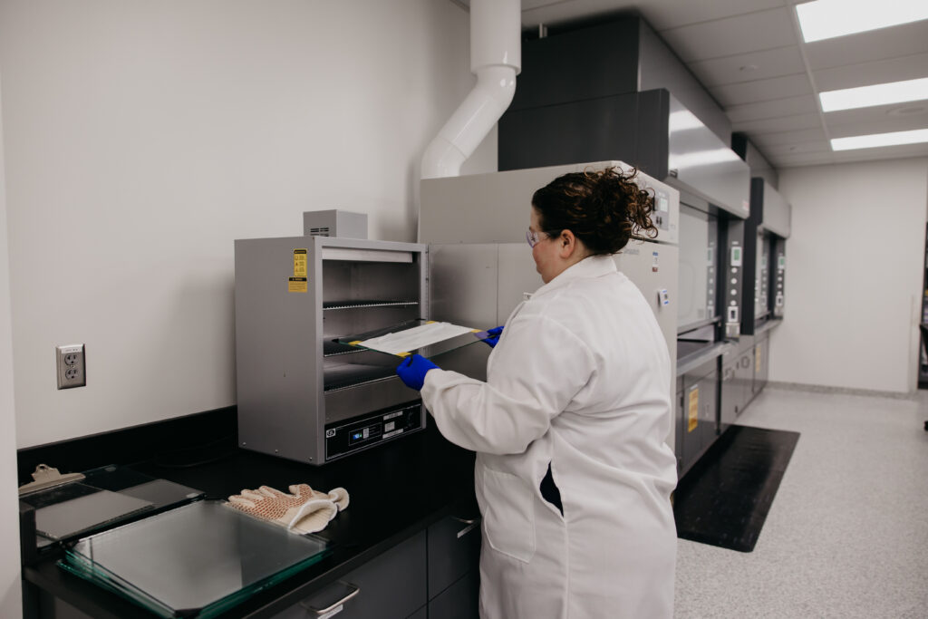 Technician in white gown operating equipment beneath an overhead exhaust hood