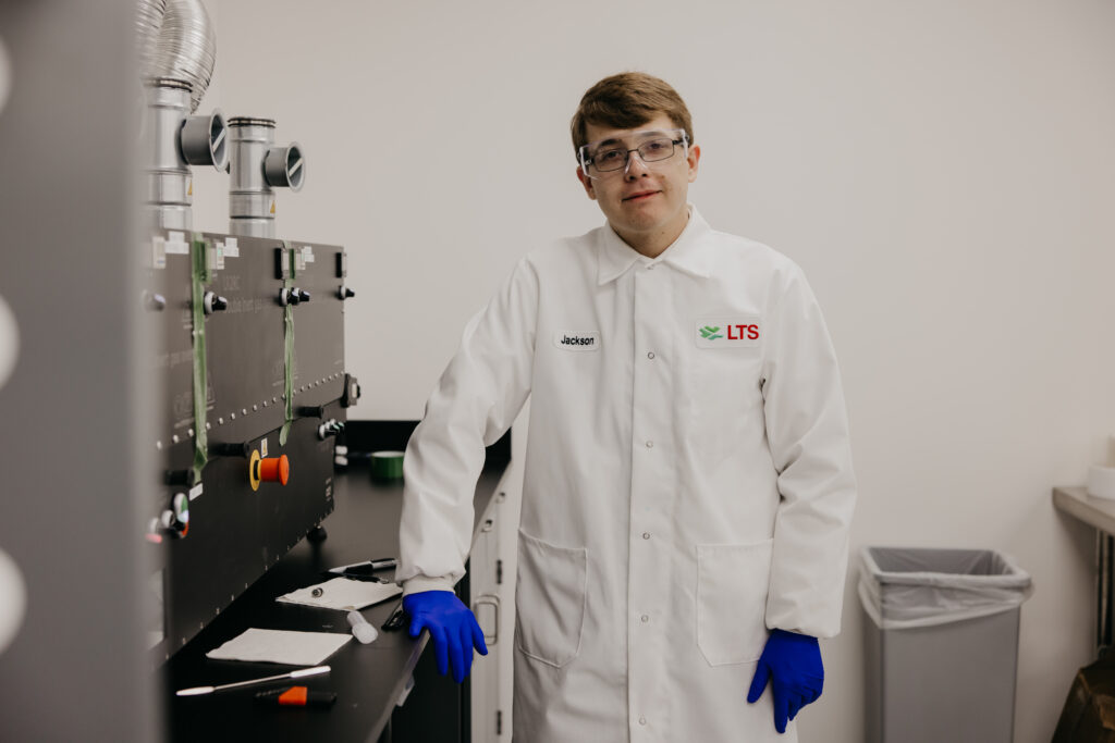 Lab technician named Jackson in white LTS coat standing beside pharmaceutical manufacturing equipment