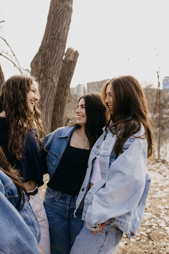 Four senior friends in denim laughing and turning toward each other near bare trees