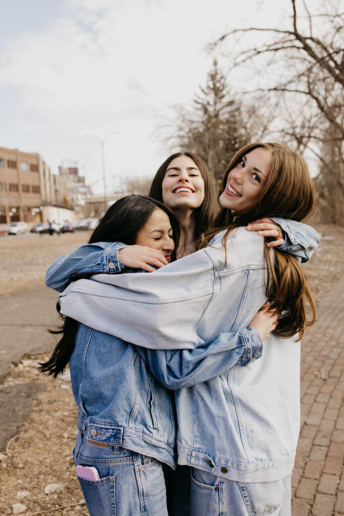 Two senior friends in denim jackets hugging tightly with bare trees and city buildings in the background