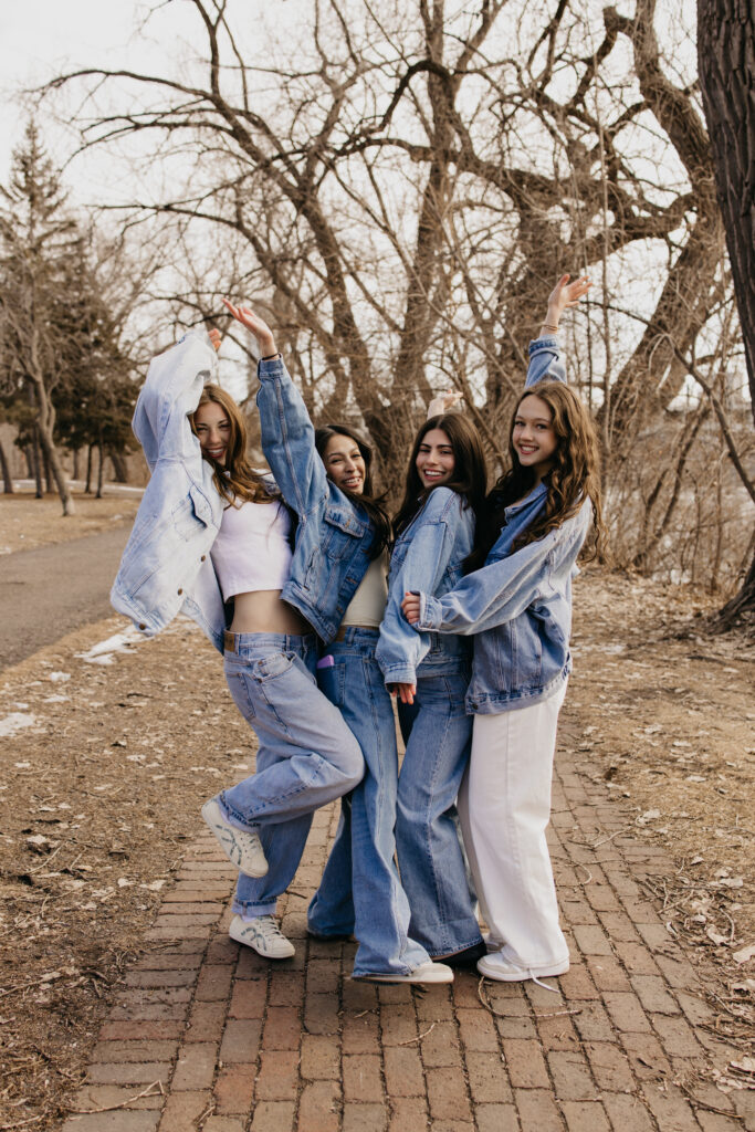 Four senior friends in denim jumping and posing playfully on a brick path lined with bare trees
