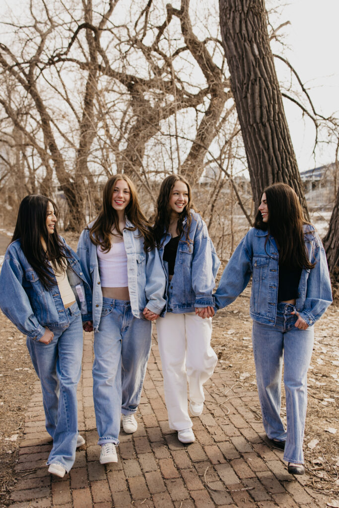 Four senior friends in matching denim jackets walking hand in hand under a bare tree on a brick path