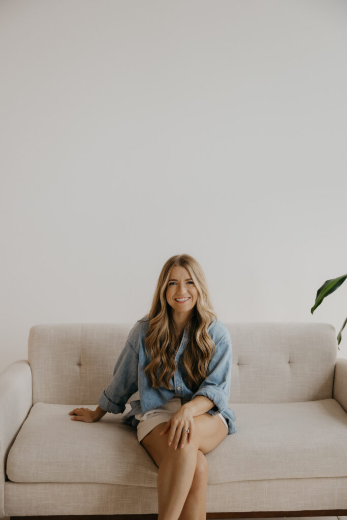 Content creator sitting on couch in denim shirt smiling with legs crossed