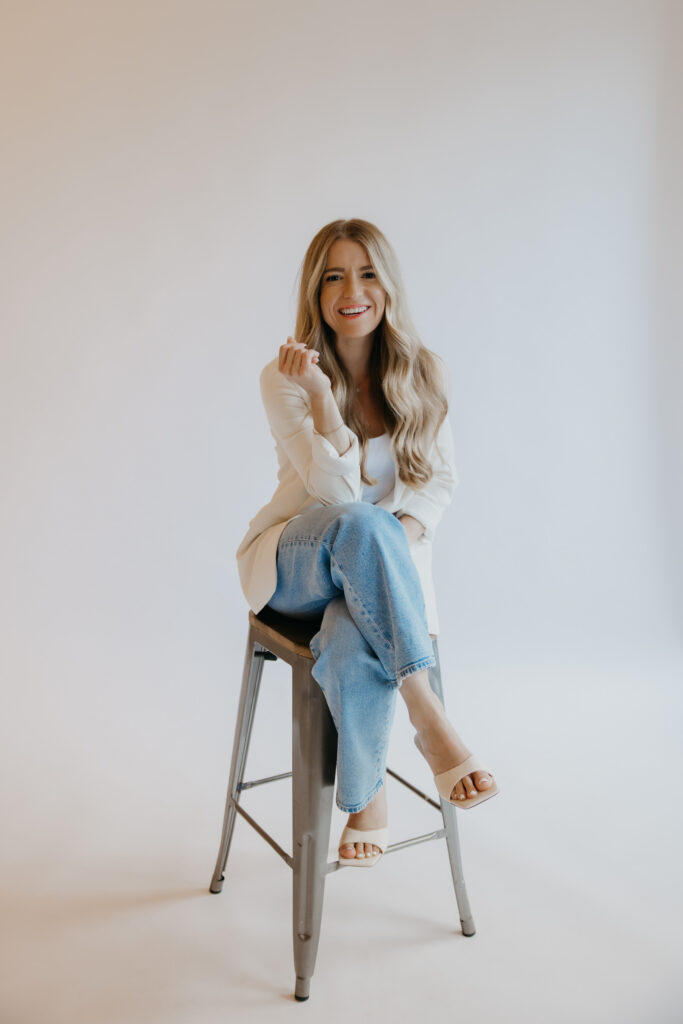 Content creator perched on stool in cream cardigan and jeans, hand raised