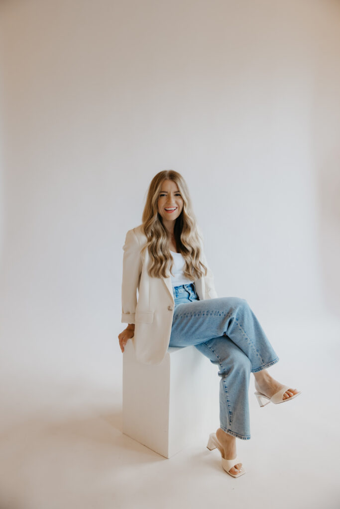 Content creator seated on white block in cream blazer and jeans, three-quarter view