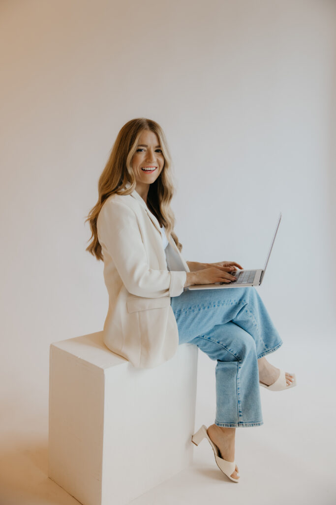 Content creator seated on white block typing on laptop in cream blazer