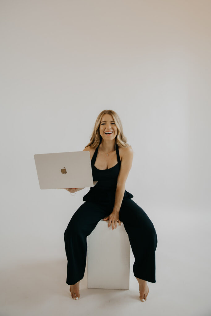 Content creator seated on white block laughing while holding laptop, black dress