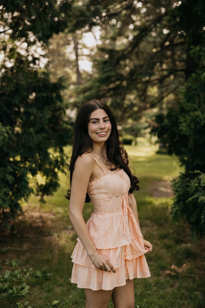 Senior in peach ruffled dress smiling while walking through a tree-lined park