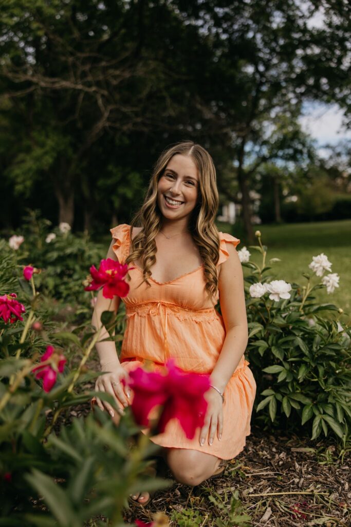 Senior in orange dress kneeling beside magenta and white peony blooms, smiling