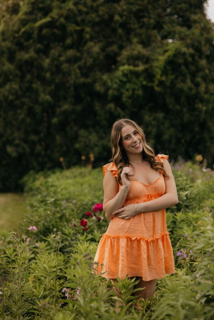Senior in orange dress standing among magenta flowers, hand near her hair