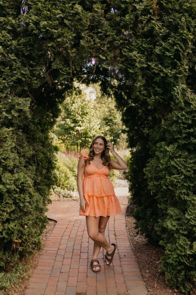 Senior in orange dress walking a brick path through a garden archway, touching her hair