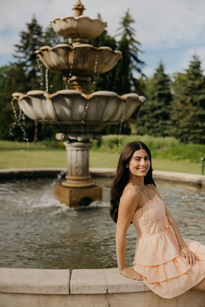 Senior in peach dress sitting on the edge of a tiered garden fountain, smiling