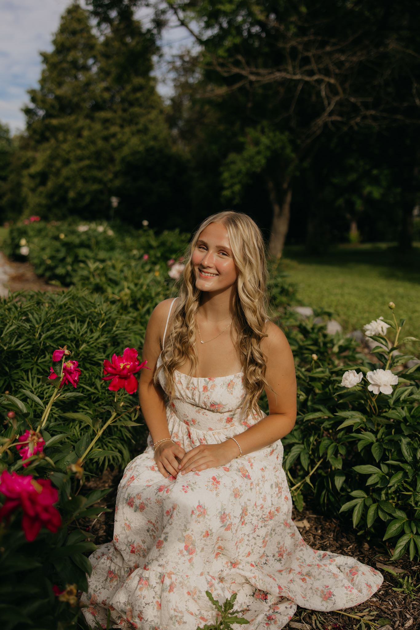 Senior girl in a floral dress sitting among pink and white peonies during a personalized senior photoshoot in a garden