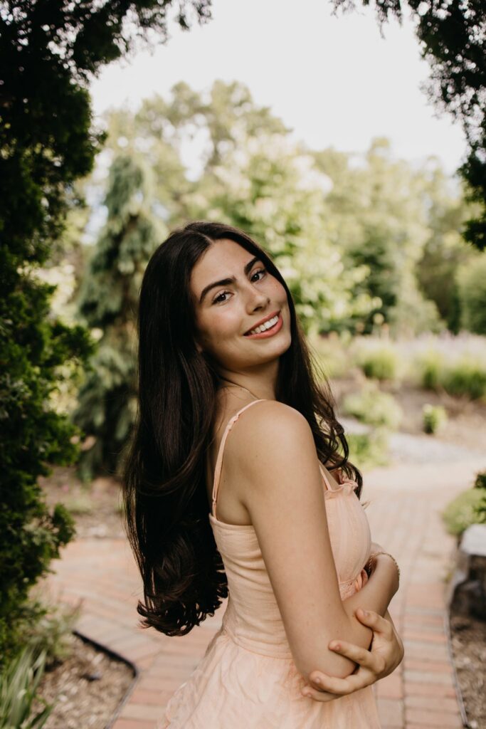 Dark-haired senior in peach dress looking over her shoulder on a brick garden path
