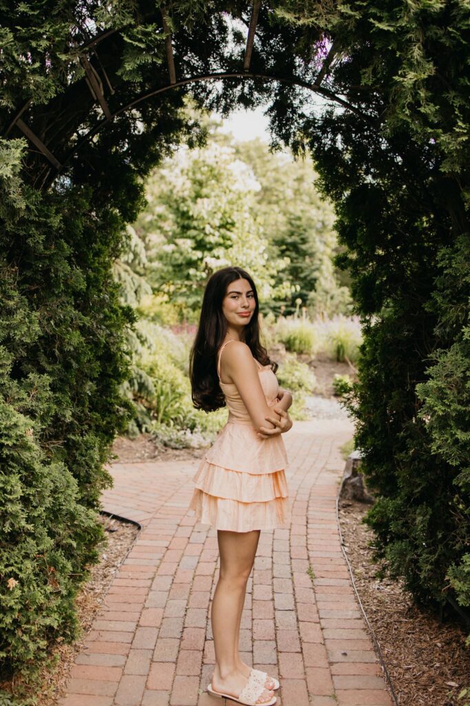 Senior in peach dress walking a brick path through a garden archway