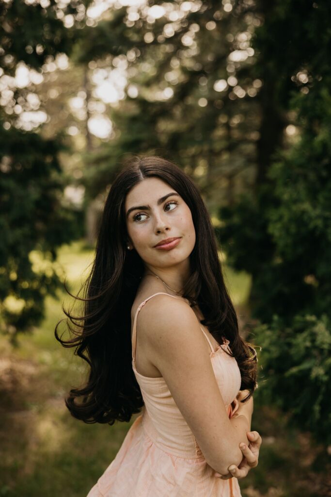 Dark-haired senior in peach dress glancing over her shoulder with hair blowing in evergreen backdrop