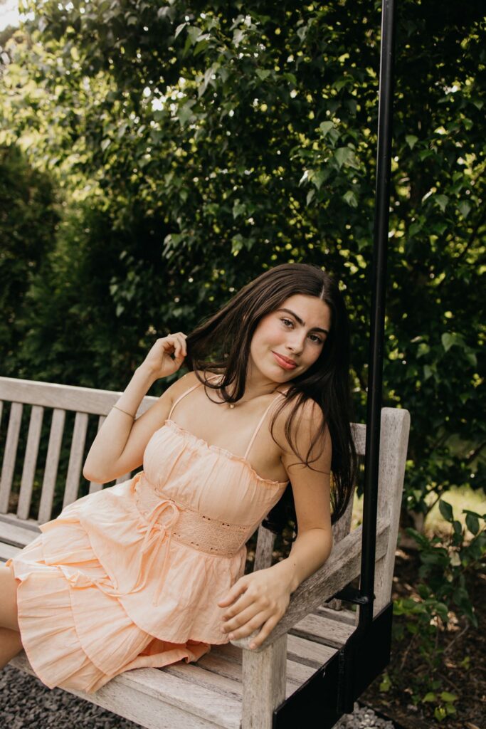Close-up of senior in peach dress on a bench swing, hand raised on a post